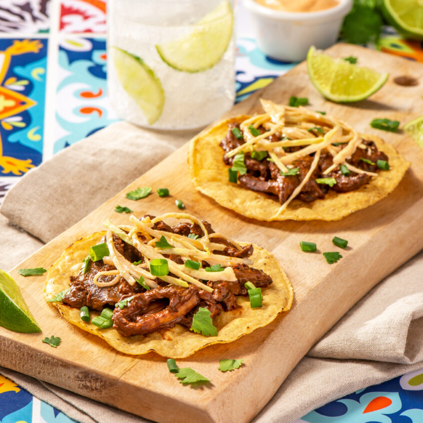 Two corn tostadas topped with mushrooms infused in mole, cabbage, and green onions, placed on a wooden plank.